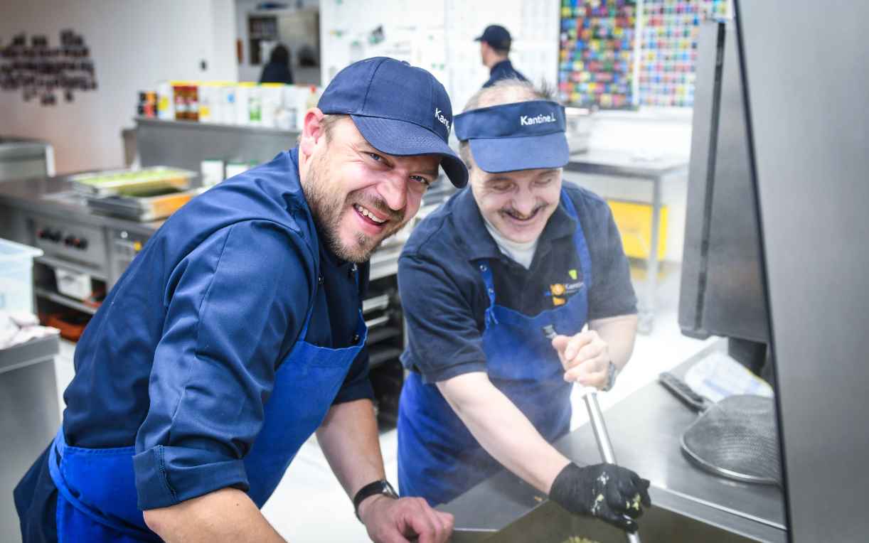 Zwei Köche lächeln in einer modernen Küche, während sie gemeinsam an einem großen Kochtopf arbeiten. Die Atmosphäre ist freundlich und einladend, jeder trägt eine Uniform mit Schürze. Die Küche ist gut ausgestattet mit verschiedenen Utensilien und Geräten.