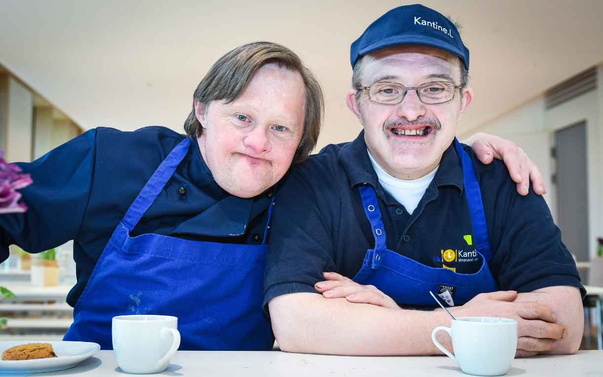 Zwei Männer in blauen Kochschürzen lächeln fröhlich in die Kamera. Sie sitzen an einem Tisch mit Tassen und einem Dessert. Der größere Mann hat eine Brille und trägt eine Mütze, während der andere eine lockerere Frisur hat. Beide vermitteln ein Gefühl von Freundschaft und Freude.