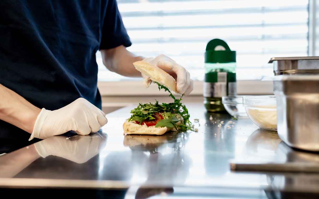 Ein Mensch in weißen Handschuhen bereitet ein Sandwich vor. Auf einer Edelstahloberfläche werden frische Zutaten wie Salat und Tomaten in ein aufgeschnittenes Brot gelegt. Im Hintergrund sind Küchenutensilien und eine Flasche Öl sichtbar.