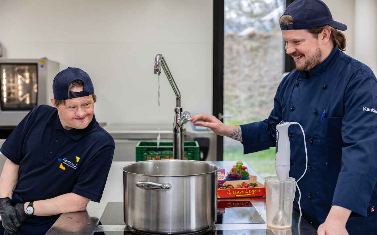 Zwei Personen in einer Küchenumgebung: Ein Mann trägt eine blaue Uniform und steht am Tisch neben einem großen Kochtopf, während der andere Mann, ebenfalls in Kochkleidung, Wasser aus einem Wasserhahn in den Topf gießt. Beide wirken erfreut und lachen.