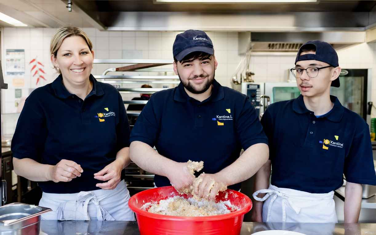 Drei Personen stehen in einer Küche. Eine Frau lächelt, während ein Mann mit Händen in einer großen roten Schüssel arbeitet. Ein junger Mann schaut konzentriert. Alle tragen dunkle Poloshirts und Schürzen. Im Hintergrund sind Küchengeräte und Regale zu sehen.