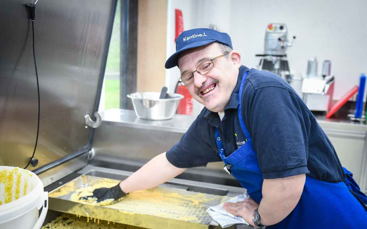 Ein Mensch mit Brille und einer blauen Arbeitskleidung lächelt, während er mit einer Hand eine Oberfläche in einer Küchenumgebung reinigt. Im Hintergrund sind Küchengeräte und Utensilien sichtbar.
