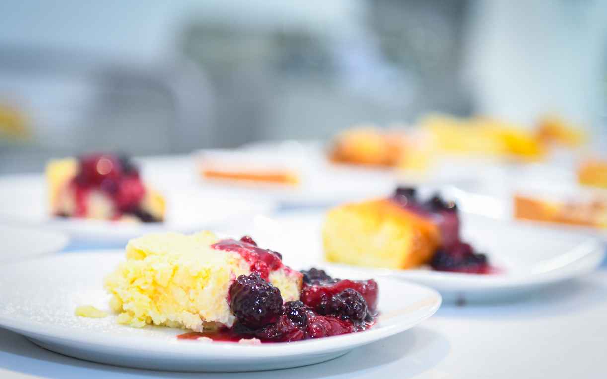 Ein Teller mit einem Stück Käsekuchen, garniert mit einer Vielzahl von Beeren und einer fruchtigen Sauce. Im Hintergrund sind weitere Teller mit ähnlichen Desserts sichtbar. Die Farben sind lebendig und die Anrichtung sieht appetitlich aus.