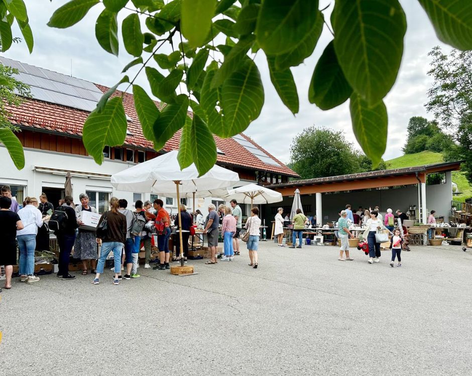 Auf dem Bild ist eine belebte Marktszene vor einem Gebäude zu sehen. Menschen stehen in Gruppen, manche unter Sonnenschirmen, während sie miteinander sprechen oder stöbern. Es gibt Tische mit verschiedenen Artikeln zum Verkauf. Bäume und eine grüne Umgebung sind im Hintergrund sichtbar.