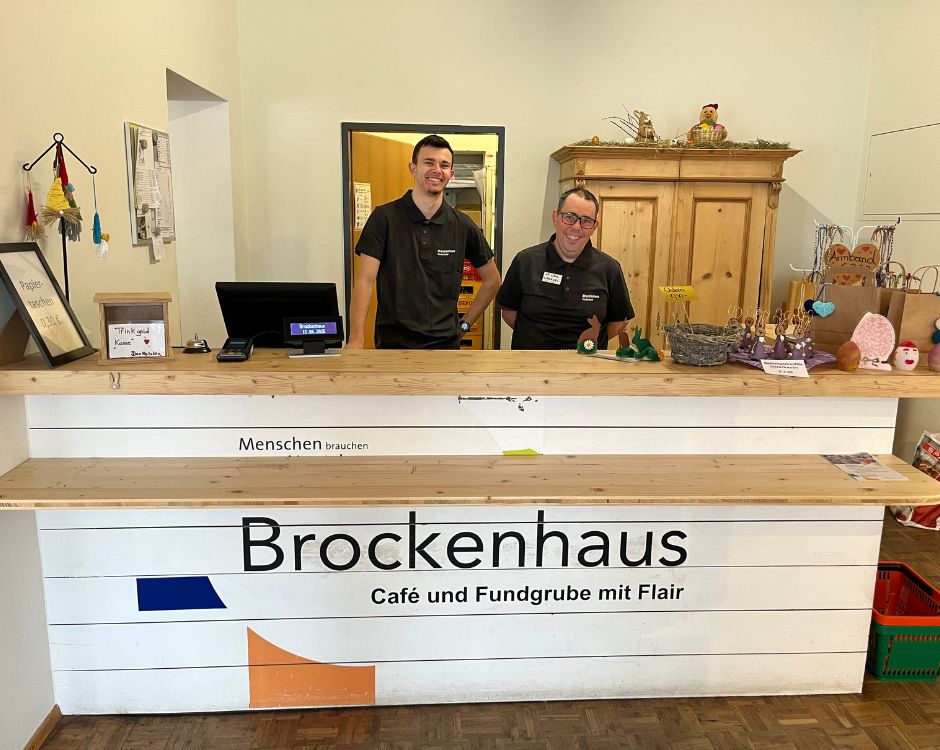 Alt-Text: Empfangstresen im Brockenhaus mit zwei Mitarbeitern. Der Tresen ist aus Holz, mit der Aufschrift „Brockenhaus Café und Fundgrube mit Flair“. Im Hintergrund sind verschiedene Dekorationsartikel und ein Kassensystem zu sehen.
