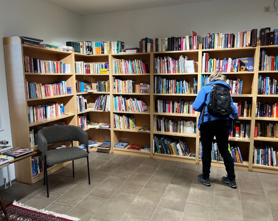 Eine Person mit einem Rucksack steht vor einem Bücherregal in einer Bibliothek und betrachtet die Bücher. Rechts und links sind mehrere Regale mit einer Vielzahl von Büchern zu sehen. Ein leerer Stuhl steht in der Nähe.