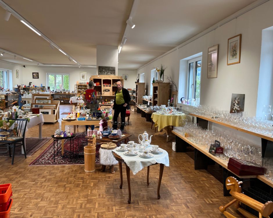 Ein gepfleckter Raum mit Tischen voller Geschirr, Gläsern und Dekorationsartikeln. Zwei Personen sind im Hintergrund aktiv. Regalwände sind mit weiteren Gegenständen bestückt. Der Boden ist mit Holzdielen ausgelegt, und große Fenster sorgen für Beleuchtung.