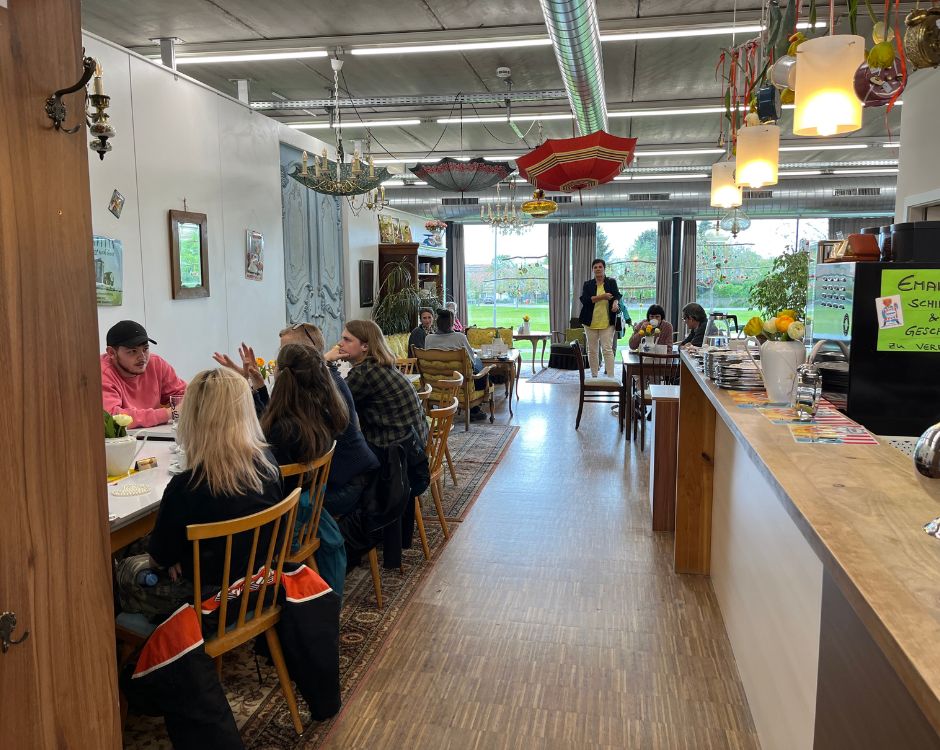 Ein offener Raum mit einer langen Tafel, an der mehrere Menschen sitzen und miteinander reden. Im Hintergrund ist eine Theke mit Lebensmitteln und Getränken sowie Dekorationselementen wie Regenschirmen zu sehen. Durch große Fenster strömt Tageslicht herein.