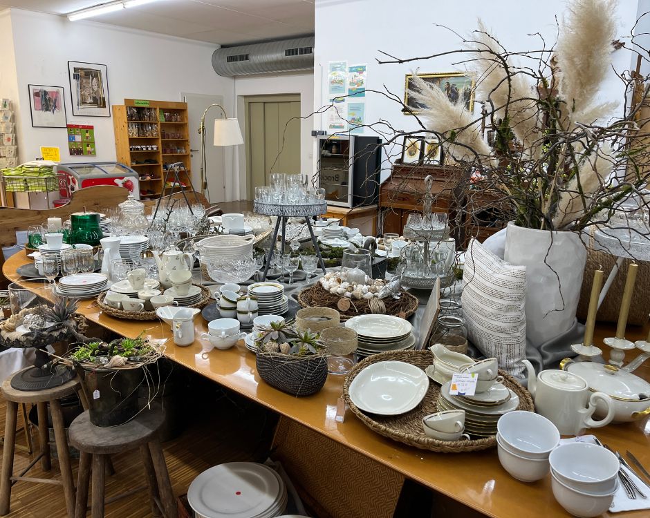 Ein einladender Tisch mit einer Vielzahl von Geschirr, Glaswaren und Dekoartikeln. Es gibt Tassen, Teller, Vasen und Pflanzenarrangements, die zusammen eine gemütliche Atmosphäre schaffen. Im Hintergrund sind Regale mit weiteren Haushaltsartikeln sichtbar.