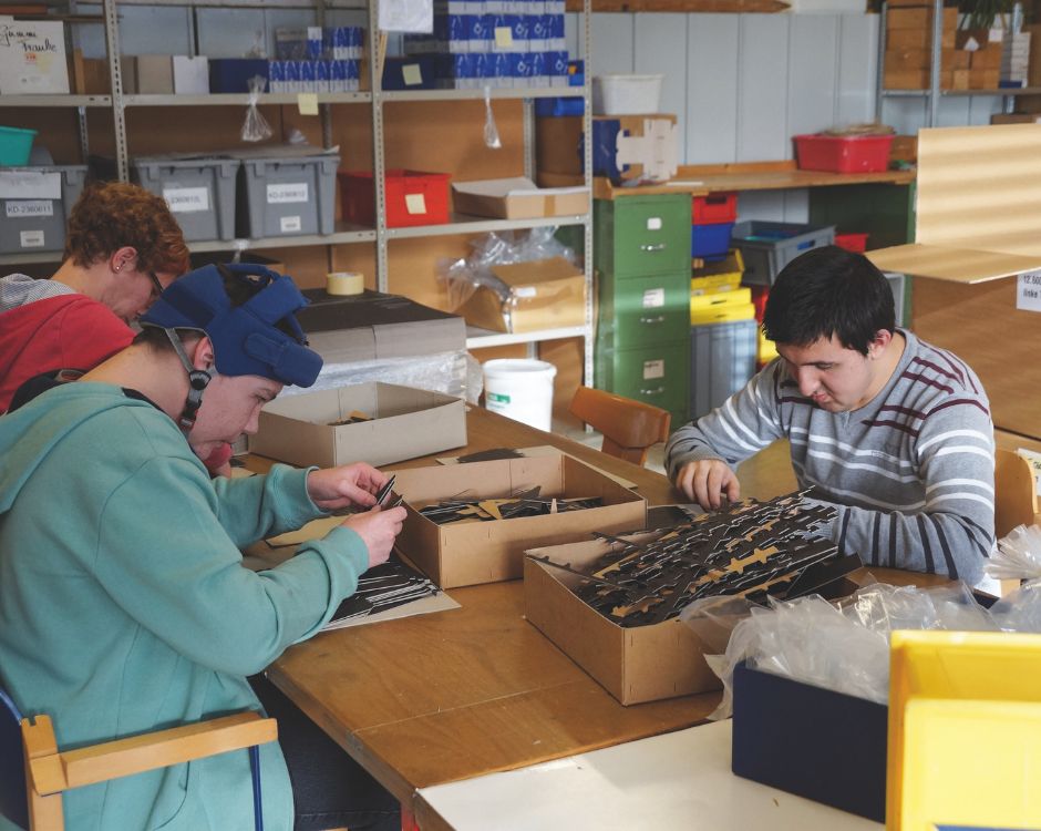 Drei Personen sitzen an einem Tisch in einem Werkraum und arbeiten an Bastelprojekten. Zwei Personen konzentrieren sich auf das Sortieren von Materialien in Kisten, während eine dritte Person im Hintergrund an einem anderen Projekt arbeitet. Der Raum ist gut beleuchtet und mit Regalen voll von Materialien ausgestattet.