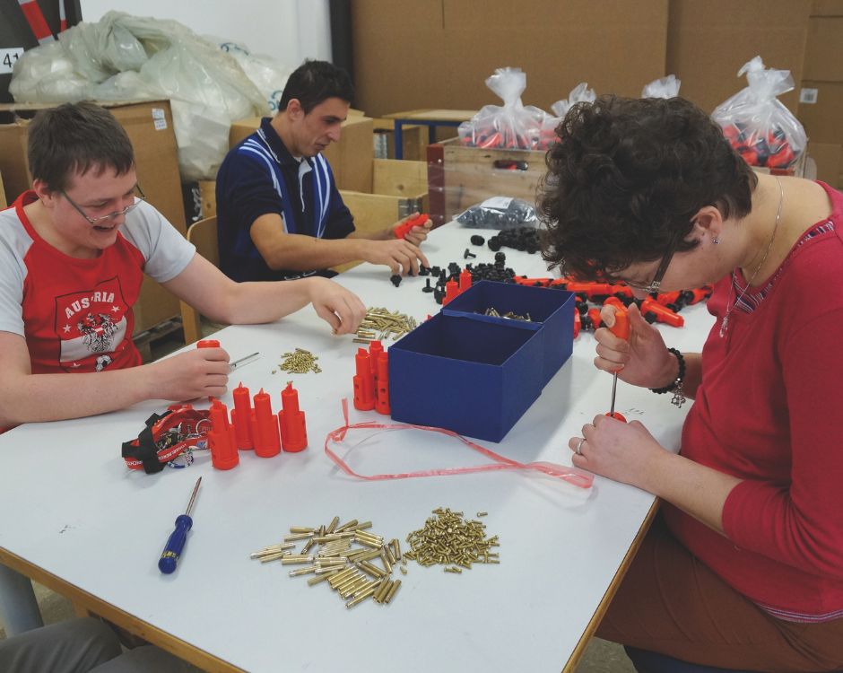 Drei Personen sitzen an einem Tisch und arbeiten an verschiedenen Aufgaben. Sie sortieren und montieren Teile, darunter orangefarbene Objekte und kleine Metallteile. Der Raum ist hell erleuchtet und im Hintergrund sind Kisten sichtbar.