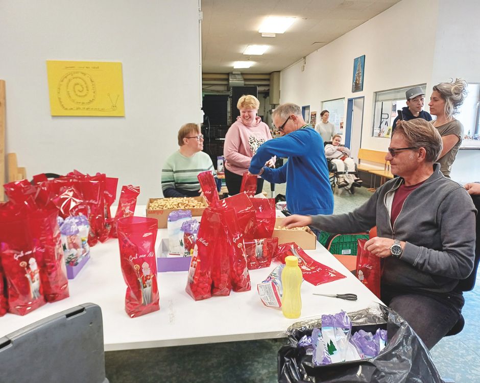 Eine Gruppe von Menschen arbeitet an einem Tisch, der mit roten Tüten und verschiedenen Lebensmitteln bedeckt ist. Im Hintergrund sitzen einige Personen auf Stühlen. Die Atmosphäre ist freundlich und einladend, mit einem Kunstwerk an der Wand.