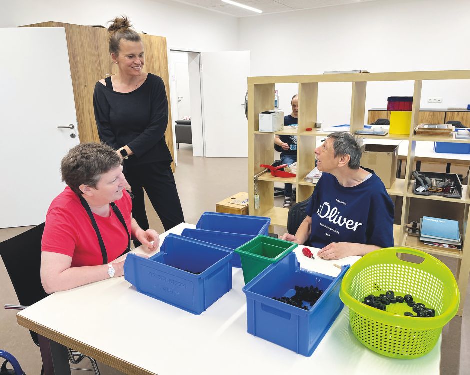 Eine Gruppe von drei Personen in einem Raum. Zwei Frauen sitzen an einem Tisch mit bunten Behältern vor sich, während eine dritte Person, die steht, sie beobachtet. Die Atmosphäre ist freundlich und kooperativ, mit einer konzentrierten Aktivität am Tisch.