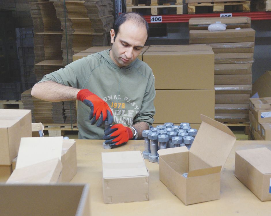 Ein Mann mit roten Handschuhen verpackt Gegenstände in einem Lagerraum. Er sitzt an einem Tisch, umgeben von leeren Kartons und fertigen Verpackungen. Im Hintergrund sind weitere Kartons gestapelt.