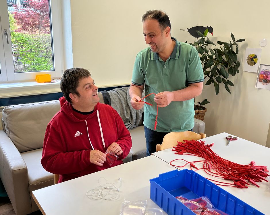 Zwei Männer sitzen an einem Tisch, auf dem rote Bänder und eine blaue Box liegen. Ein Mann im roten Hoodie lächelt, während der andere in einem grünen Poloshirt ihm ein Stück von einem Band zeigt. Im Hintergrund sind eine Couch und Pflanzen sichtbar.