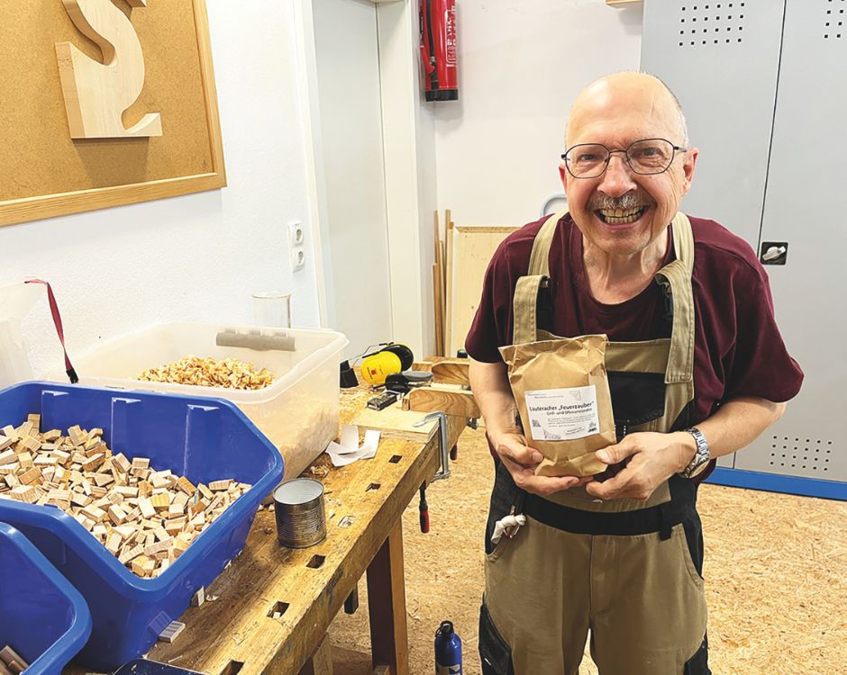Ein älterer Mann mit Brille und einem breiten Lächeln steht in einer Werkstatt. Er hält eine braune Papiertüte in den Händen und steht vor einem Tisch mit Holzstücken, Werkzeugen und einer Kiste mit weiteren kleinen Holzstücken.
