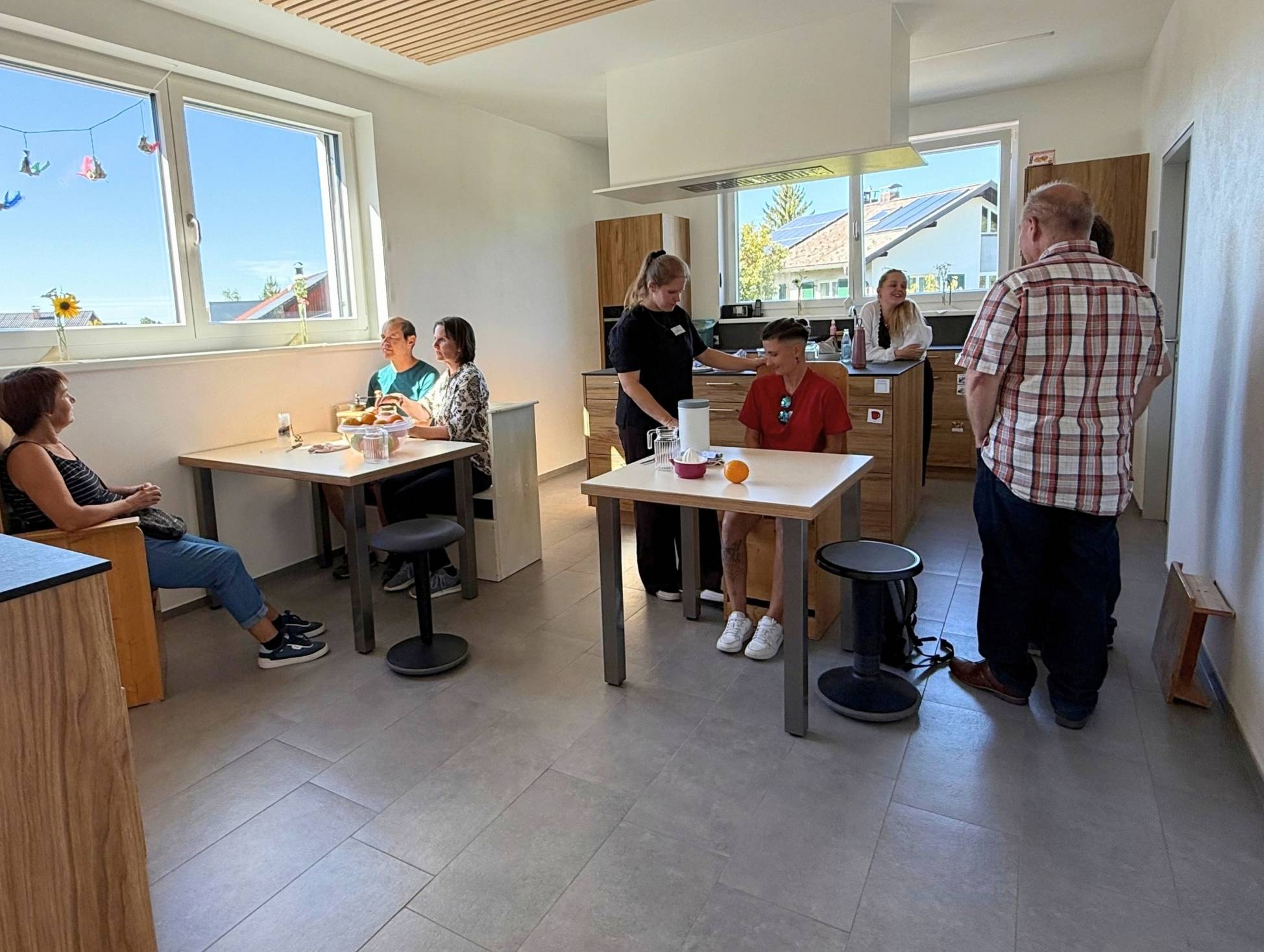 Eine helle, geräumige Küche mit mehreren Tischen. Einige Personen sitzen und unterhalten sich, während andere mit Essenszubereitung beschäftigt sind. Die Fenster zeigen einen klaren Himmel und das Außenlaufen ist sichtbar.