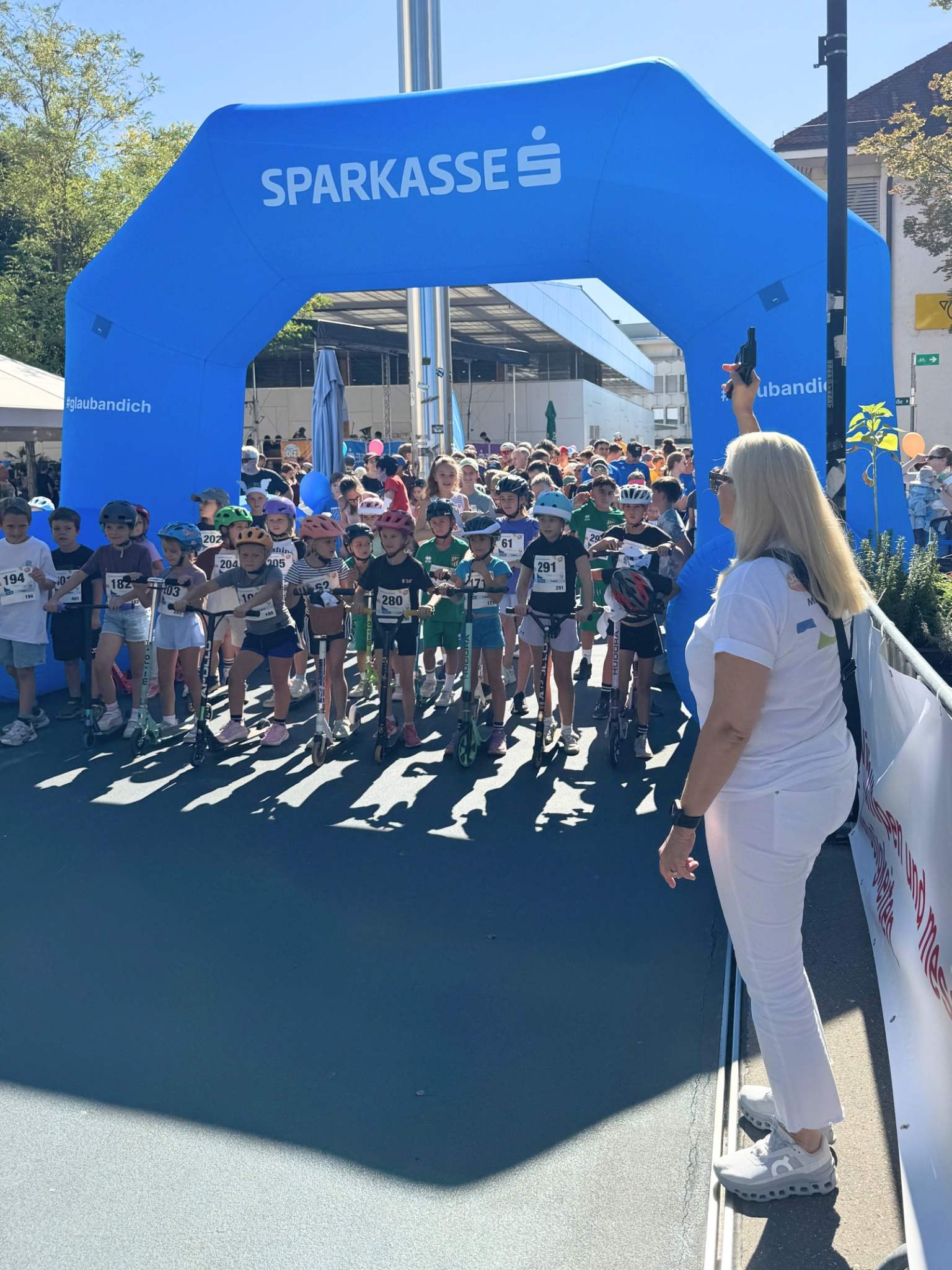 Eine Gruppe von Kindern steht an der Startlinie eines Rennens unter einem blauen Bogen mit dem Schriftzug "SPARKASSE". Eine Frau in leichter Kleidung hebt die Hand, um den Startsignal zu geben. Die Atmosphäre vermittelt Aufregung und Wettbewerb.