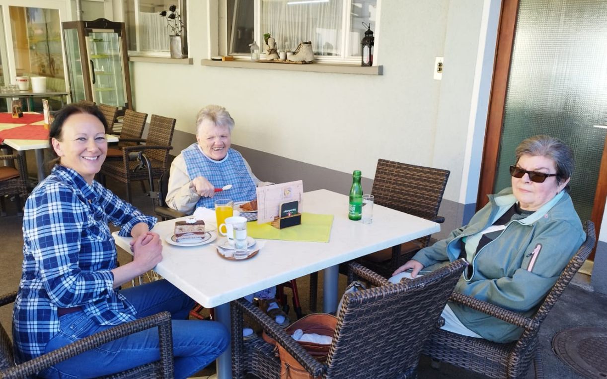 Drei Frauen sitzen an einem Tisch im Freien, ausgestattet mit Getränken und Snacks. Eine Frau lächelt in die Kamera, während die anderen beiden entspannt an ihrem Tisch sitzen. Im Hintergrund sind einige Möbel und eine Glasfront sichtbar.