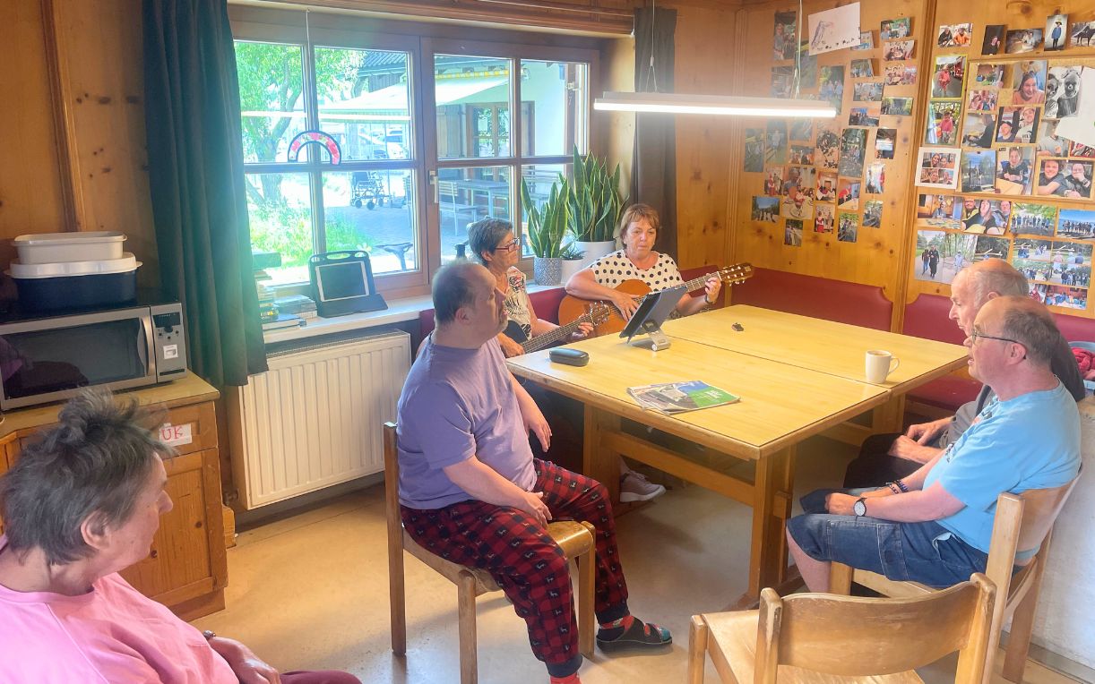 Eine Gruppe von sechs Personen sitzt in einem Raum. Eine Frau spielt Gitarre und singt, während die anderen aufmerksam zuhören. An der Wand hängen viele Fotos. Ein Tisch mit Unterlagen und Tassen steht in der Mitte des Raumes.