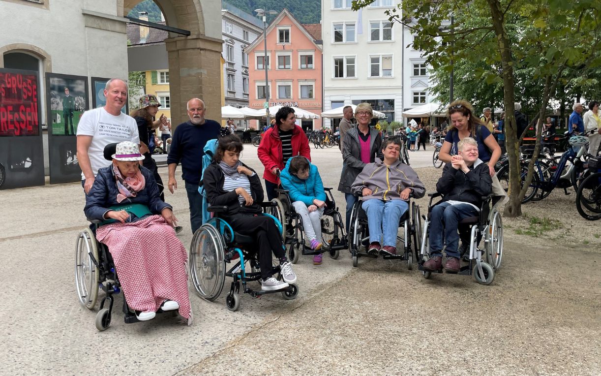Eine Gruppe von Menschen, darunter mehrere Personen im Rollstuhl, posiert in einer Stadt. Im Hintergrund sind verschiedene Gebäude und Bäume zu sehen. Die Stimmung ist freundschaftlich und einladend.