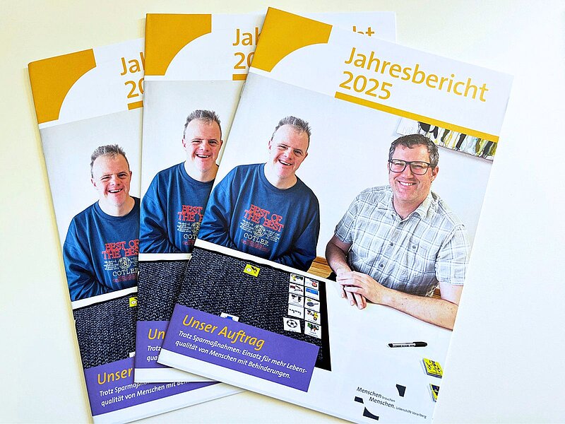 Drei Jahresberichte für 2025 liegen übereinander. Auf dem Titel sind zwei lächelnde Männer in blauen T-Shirts sowie ein dritter Mann in einem karierten Hemd abgebildet. Der Titel „Jahresbericht 2025“ und der Untertitel „Unser Auftrag“ sind klar sichtbar.