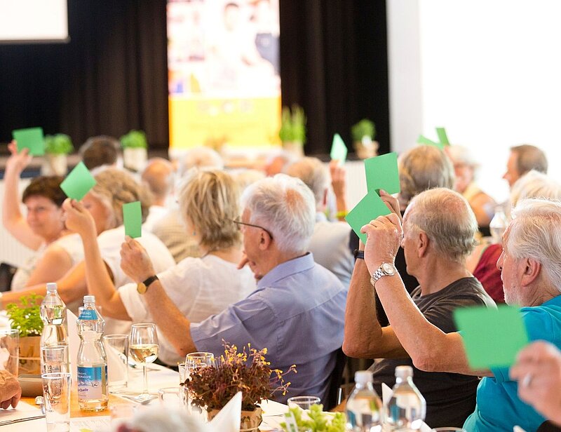 Eine Gruppe von Menschen, überwiegend Senioren, sitzt an einem gedeckten Tisch. Viele halten grüne Karten in die Höhe. Die Szenerie zeigt Teilnehmende einer Versammlung oder Abstimmung, die aktiv ihre Meinungen darstellen. Im Hintergrund ist eine Veranstaltungsbühne zu erkennen.