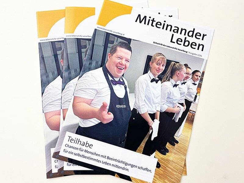 Auf dem Bild sind mehrere Stapel von Broschüren mit dem Titel "Miteinander Leben" zu sehen. Diese Broschüren behandeln Themen der Teilhabe für Menschen mit Beeinträchtigungen. Abgebildet sind auch Personen in formeller Kleidung, die lächeln und anscheinend in einem sozialen Umfeld tätig sind.