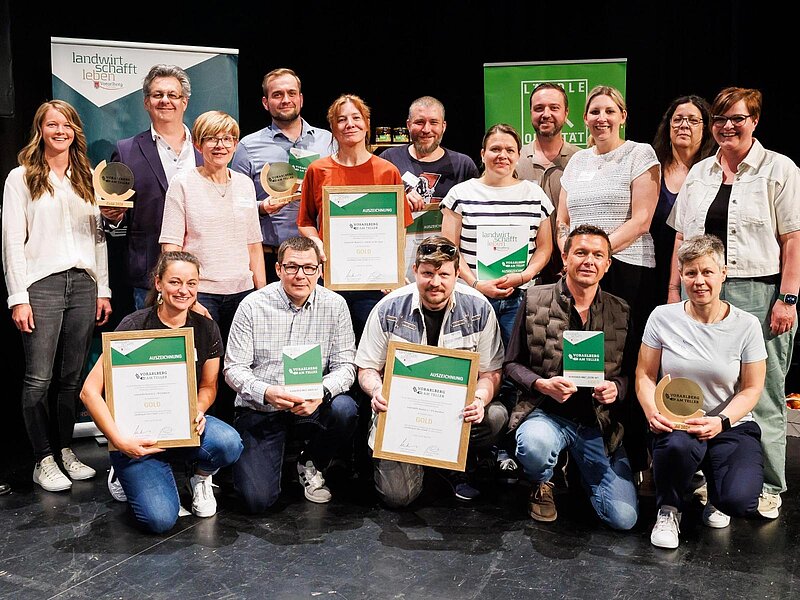 Eine Gruppe von Personen steht auf einer Bühne, umgeben von Bannern. Sie halten Urkunden und Preise in den Händen. Die Gruppe besteht aus Männern und Frauen unterschiedlichen Alters, die lächeln und stolz erscheinen.