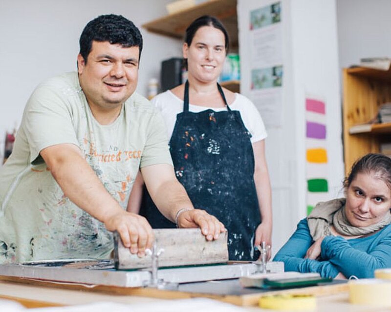 Drei Personen in einem Atelier, zwei stehen neben einer Druckmaschine. Ein Mann lächelt und dreht eine Walze, während eine Frau im Hintergrund gut sichtbar ist. Eine weitere Frau sitzt an der Seite und schaut interessiert. Der Raum wirkt kreativ und einladend.