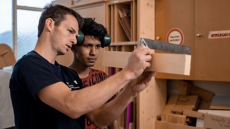 Zwei Männer stehen in einer Holzwerkstatt. Einer zeigt dem anderen, wie man ein Stück Holz mit einem Werkzeug bearbeitet. Beide sind konzentriert und schauen auf das Holzstück. Die Werkstatt ist hell und mit Holzmaterialien ausgestattet.